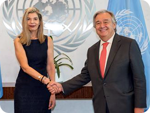 A photo of the ambassador shaking someone's hand in front of the United Nations logo.