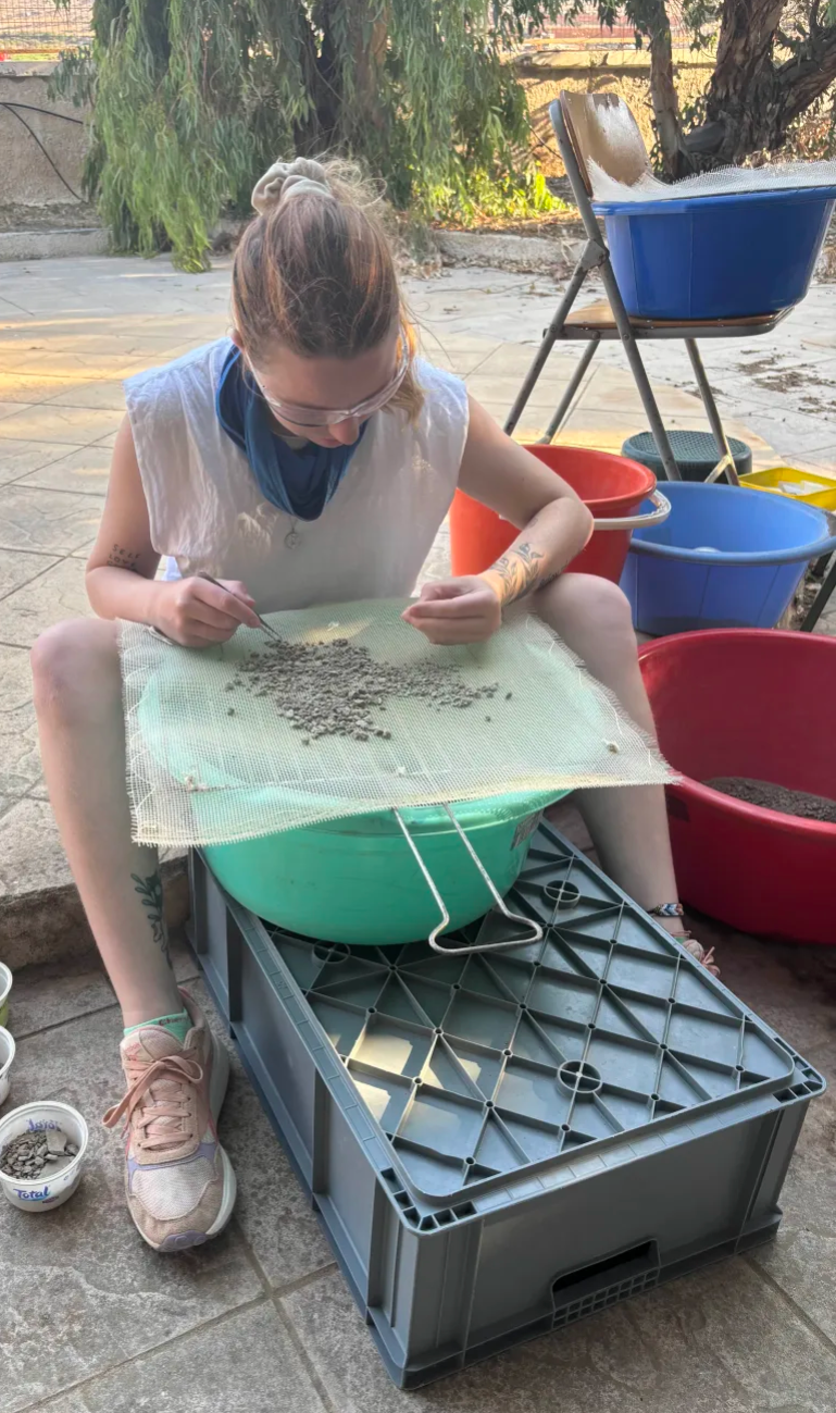 GC Sieving Photo.png - Astypalaia, Dodecanese, Greece; Gracie Carrello examining a possible find while Sieving at the lab in Maltezana.